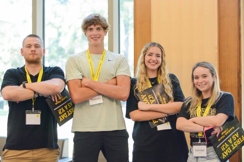Four people standing with folded arms, wearing name tags and holding yellow and black booklets, in a brightly lit room with large windows.