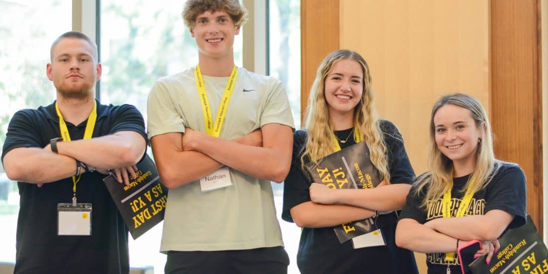 Four people standing with folded arms, wearing name tags and holding yellow and black booklets, in a brightly lit room with large windows.