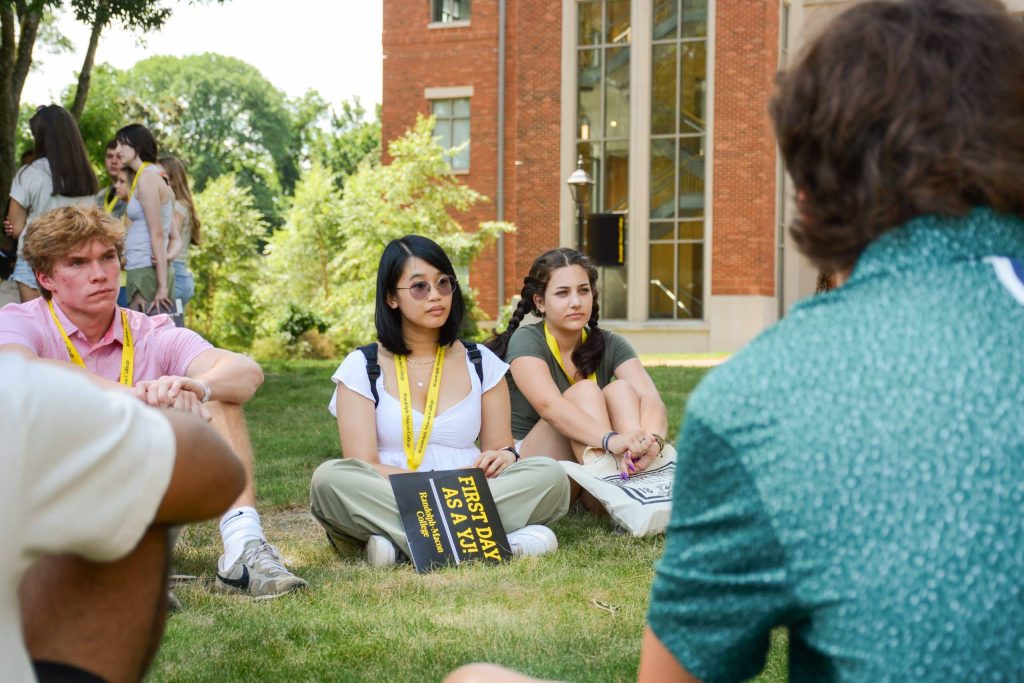 A group of students sits on grass, participating in an outdoor discussion, with a brick building in the background. One person holds a book labeled First Day.