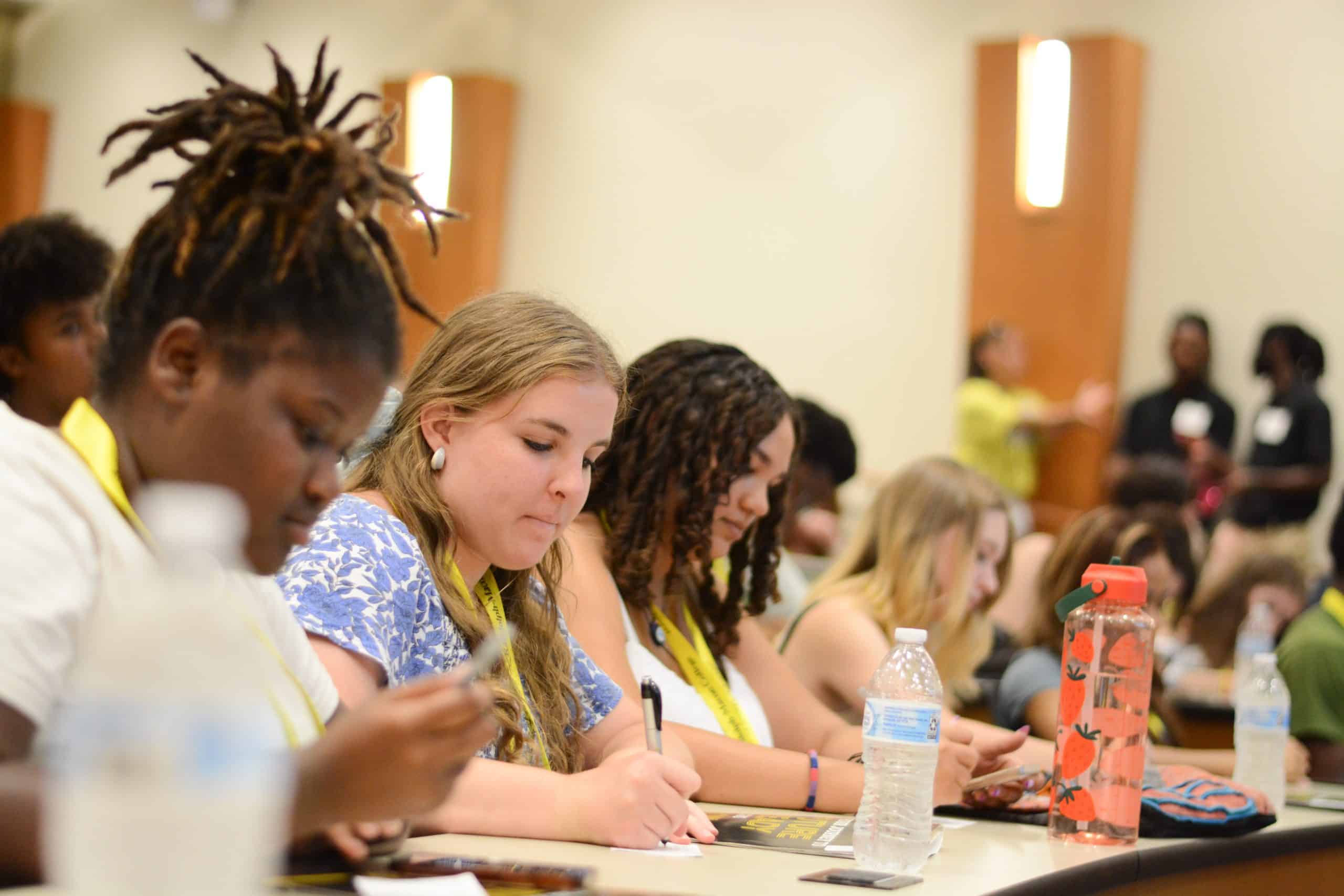 A group of students seated in a classroom, focused on writing notes. Several water bottles and papers are on the desks.