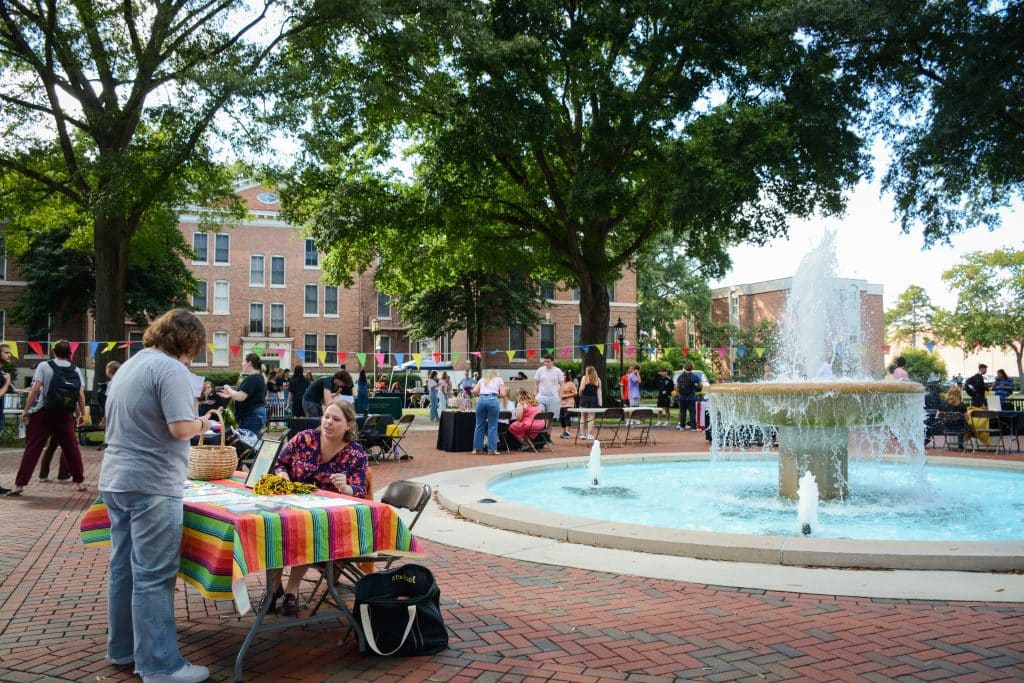 People gather around tables and a fountain at an outdoor fair with colorful bunting and trees in the background, engaging in various clubs and activities.