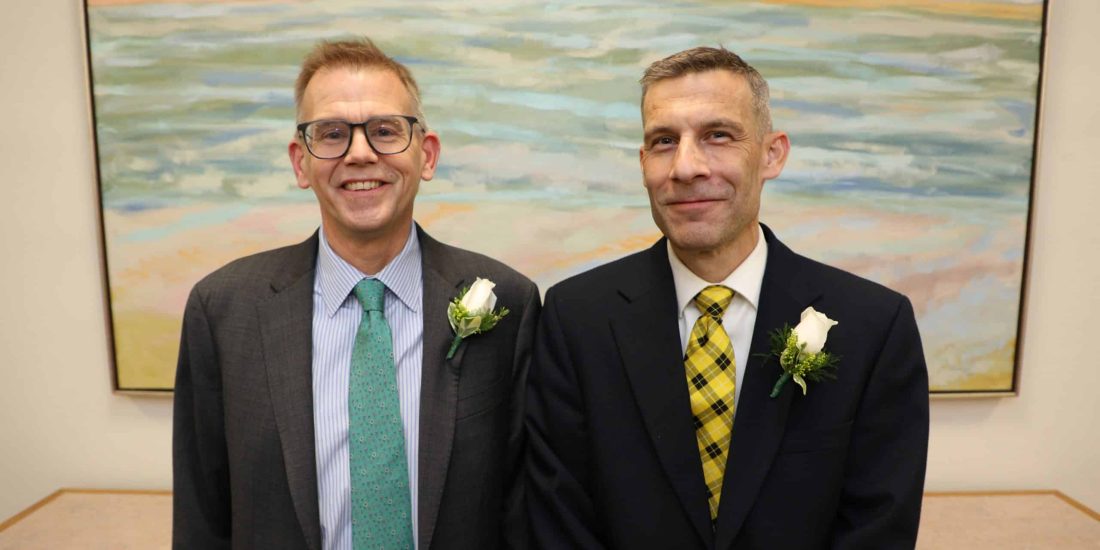 Two men in suits with white boutonnières stand in front of a seascape painting, smiling at the camera.