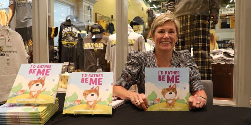 A person sitting at a table smiling, with stacks of a children's book titled "I'd Rather Be Me" displayed in front of them.