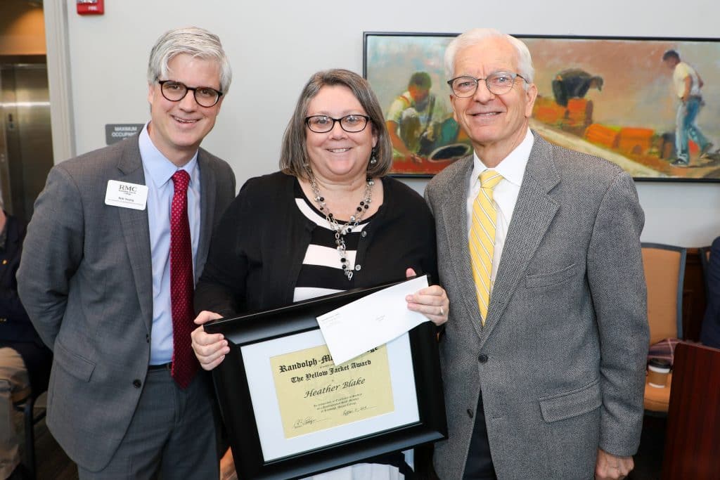 Three people stand together, smiling. The woman in the center holds a framed award and a white envelope. Two men stand on either side of her, all wearing business attire. A painting is in the background.