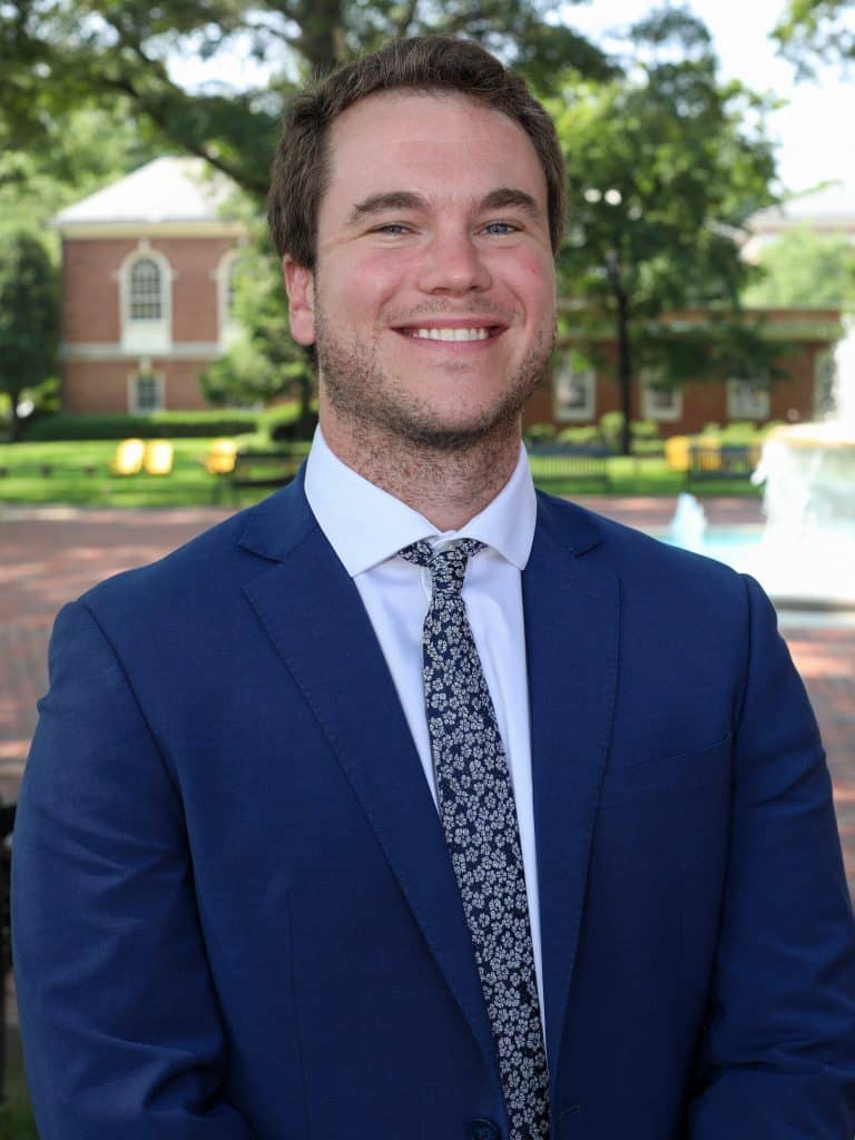 A man in a blue suit and floral tie stands outside, smiling, with a building and fountain in the background.