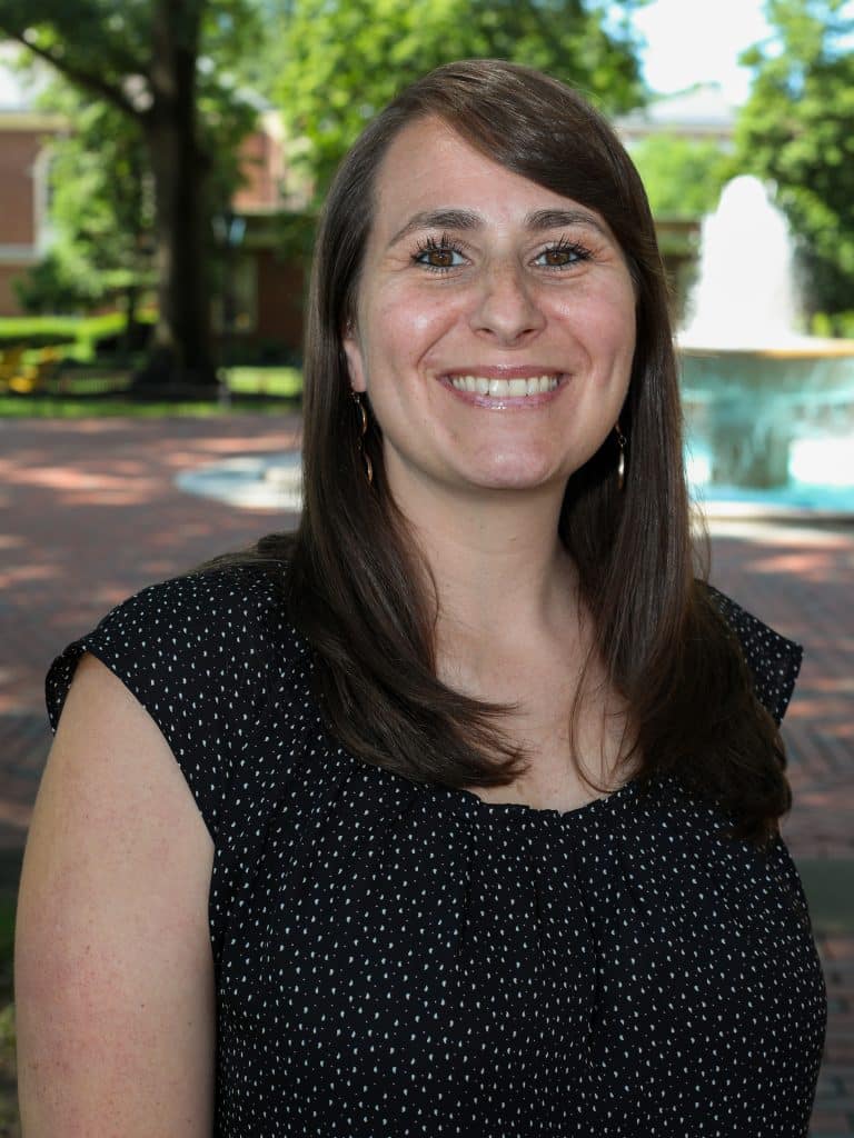 A woman with long brown hair smiles while standing in a park with a fountain and trees in the background.