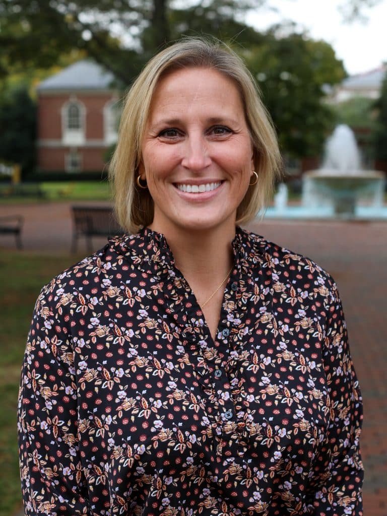 A woman with short blonde hair smiles while standing outdoors in front of a fountain and a brick building. She is wearing a floral patterned blouse.