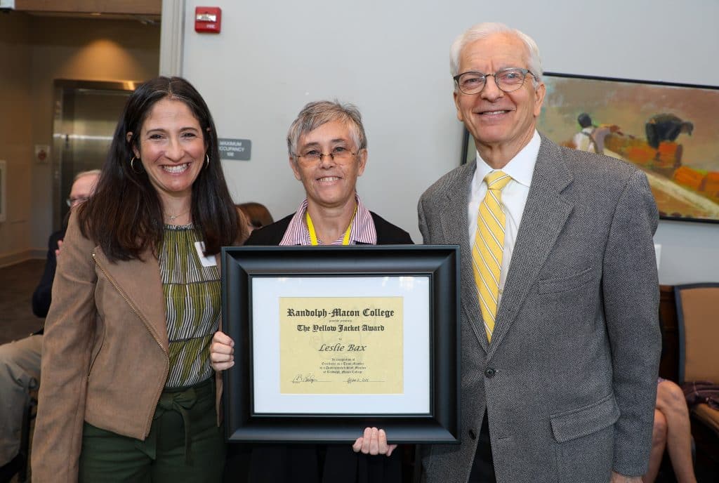 Three people stand together indoors, with the center person holding an award certificate from Randolph-Macon College.