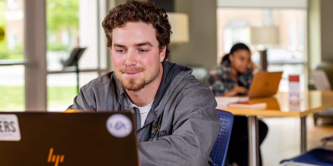 A student works at a laptop in the library with other student visible in the background.