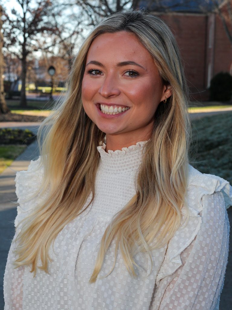 A woman with long blonde hair smiles, wearing a white blouse, standing outside.