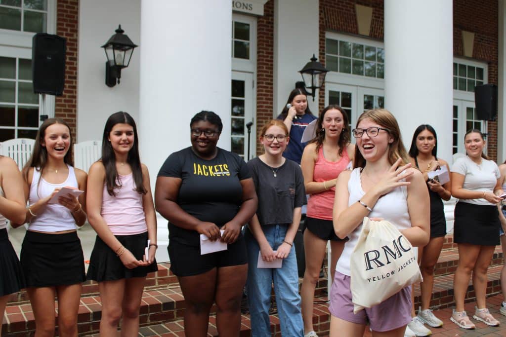 A group of people stands outside a building with columns, smiling and holding items. One person holds a bag with "RMC" on it, showcasing their participation in various clubs and activities.