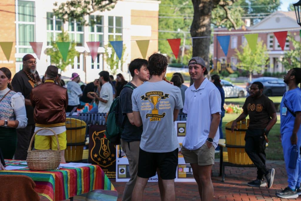 A group of people socializes at an outdoor event with colorful pennant banners, booths, and seating. A man in a gray T-shirt and two others converse in the foreground, discussing school clubs and activities.