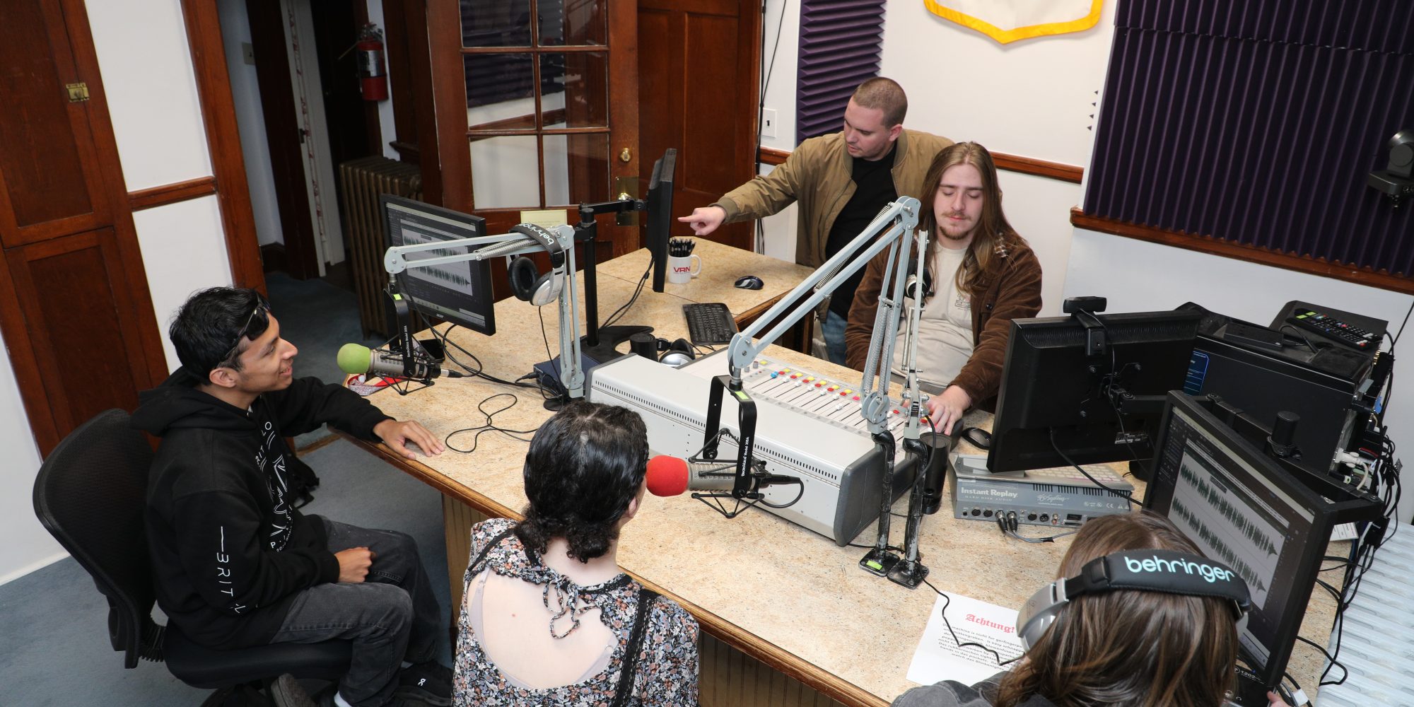 Four people are in a radio station studio engaged in communication. Two are seated at microphones, while two are standing, one adjusting audio equipment. The room, essential for broadcast studies, features soundproofing panels and multiple computer monitors.
