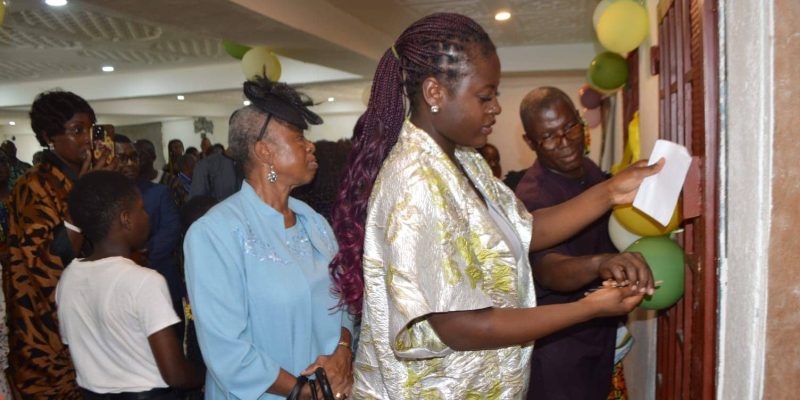 People attending an event, with a woman in a silver outfit tying balloons to a door while others watch.