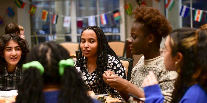 A group of people sitting around a table engaged in conversation, with international flags hanging in the background.