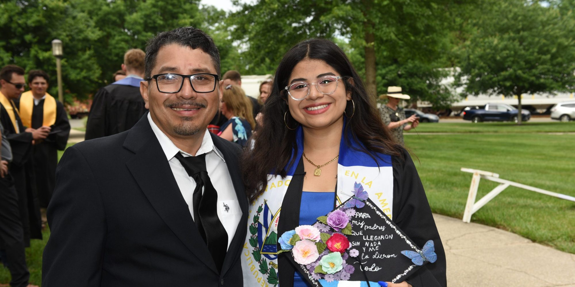 A graduate in cap and gown stands with a man outdoors, celebrating her achievements in Hispanic Literature. She holds a decorated mortarboard adorned with flowers and butterflies, symbolizing the vibrant beauty of Hispanic culture.