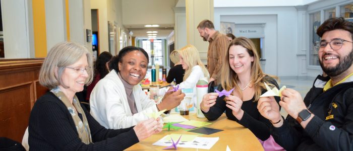 A group of people sitting at a table in a bright room, smiling and folding colorful origami paper cranes.
