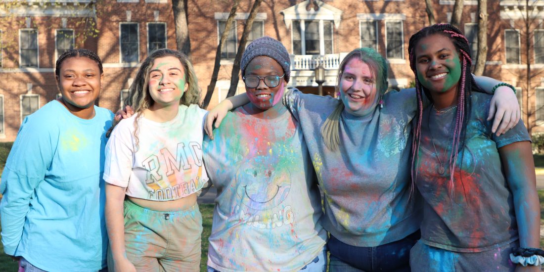 Five people stand outdoors covered in colored powder, smiling at the camera. A large brick building is in the background.
