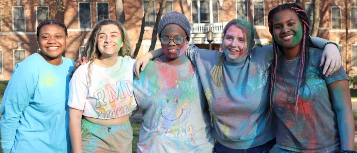 Five people stand outdoors covered in colored powder, smiling at the camera. A large brick building is in the background.