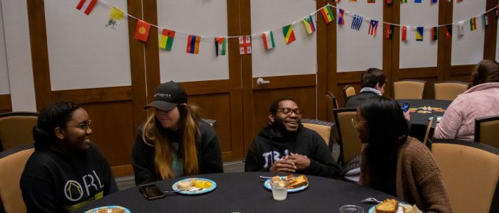 Four people sit at a round table with food and drinks. International flags decorate the background.