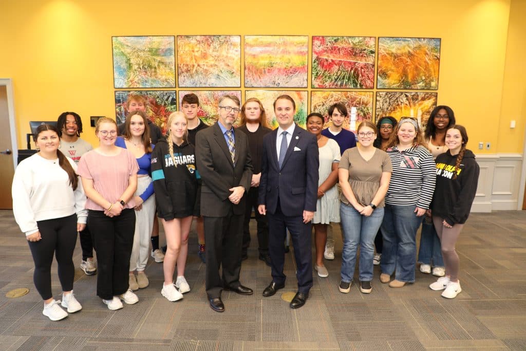 A group of people, including two men in suits, pose for a photo in front of a colorful wall art.