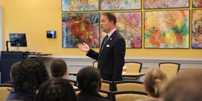 A man in a suit speaks to an audience in a room with colorful abstract art on the walls.