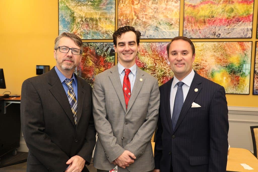 Three men in suits standing together in a room with colorful abstract artwork on the wall behind them.