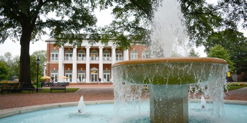 A large stone fountain with cascading water graces the entrance of a red-brick building with white columns, surrounded by trees—a peaceful spot where Virginia students often gather, discussing increased financial aid opportunities.