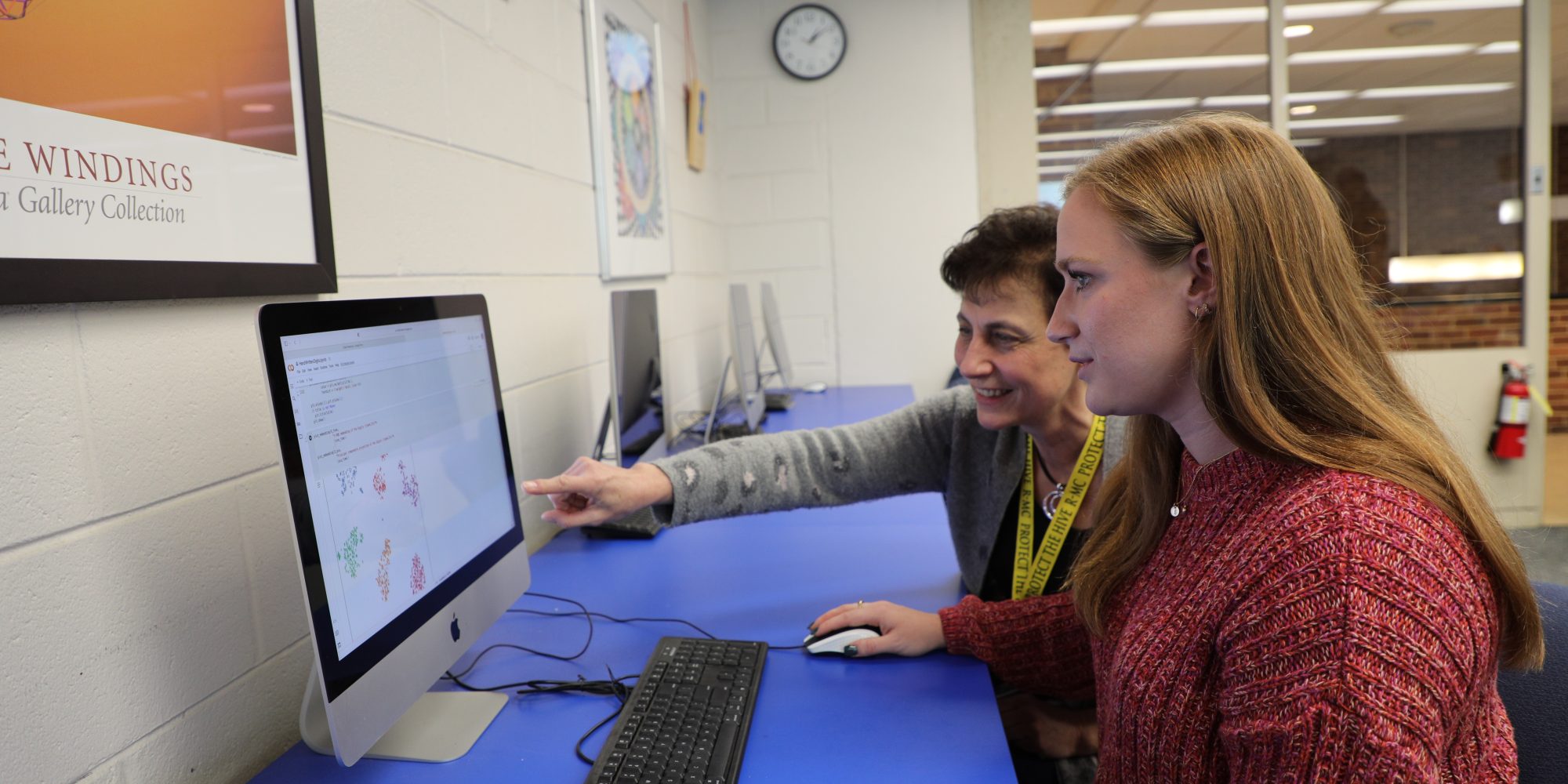A professor and student sit at a desk in front of a computer. An older person is pointing at the screen, discussing data science, while a younger person watches attentively. They are in a room filled with several computers and artworks.