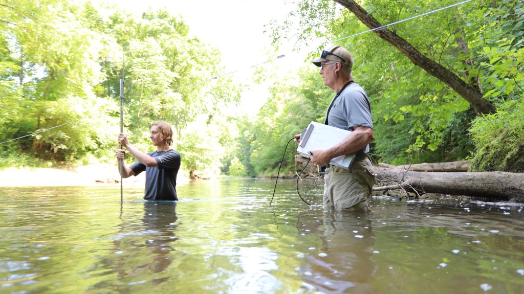 While an Environmental Studies faculty member looks on, an RMC Environmental Studies major standing in a river surrounded by trees, holding a measuring rod.