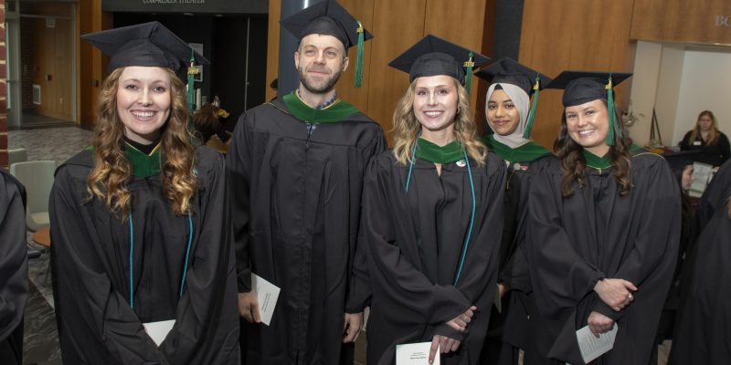 Five graduates in black gowns and caps smile, holding programs, indoors.