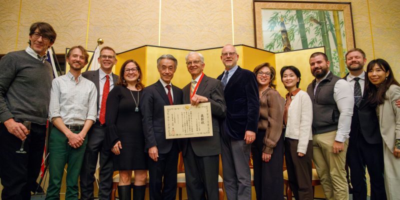 A group of people, including two men holding a certificate, pose together indoors with a decorative screen and art in the background.