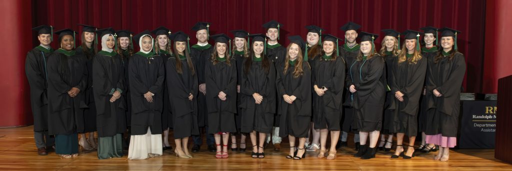 A group of graduates in caps and gowns stand on a stage against a red curtain.