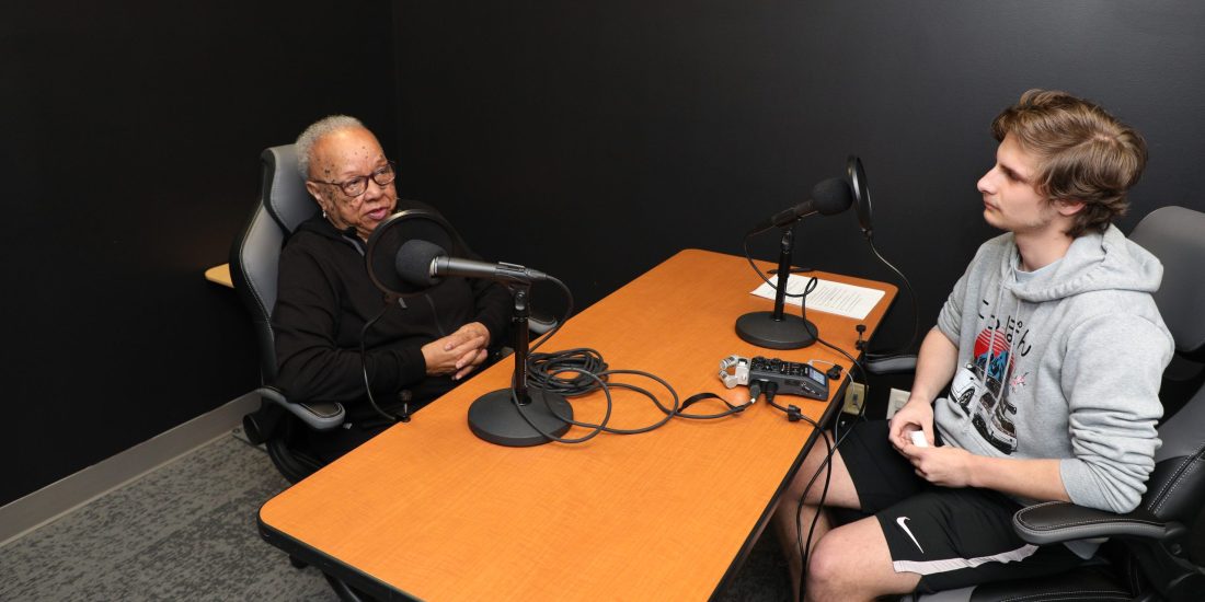 Two people sitting at a table with microphones, engaged in a conversation, in a dimly lit room.