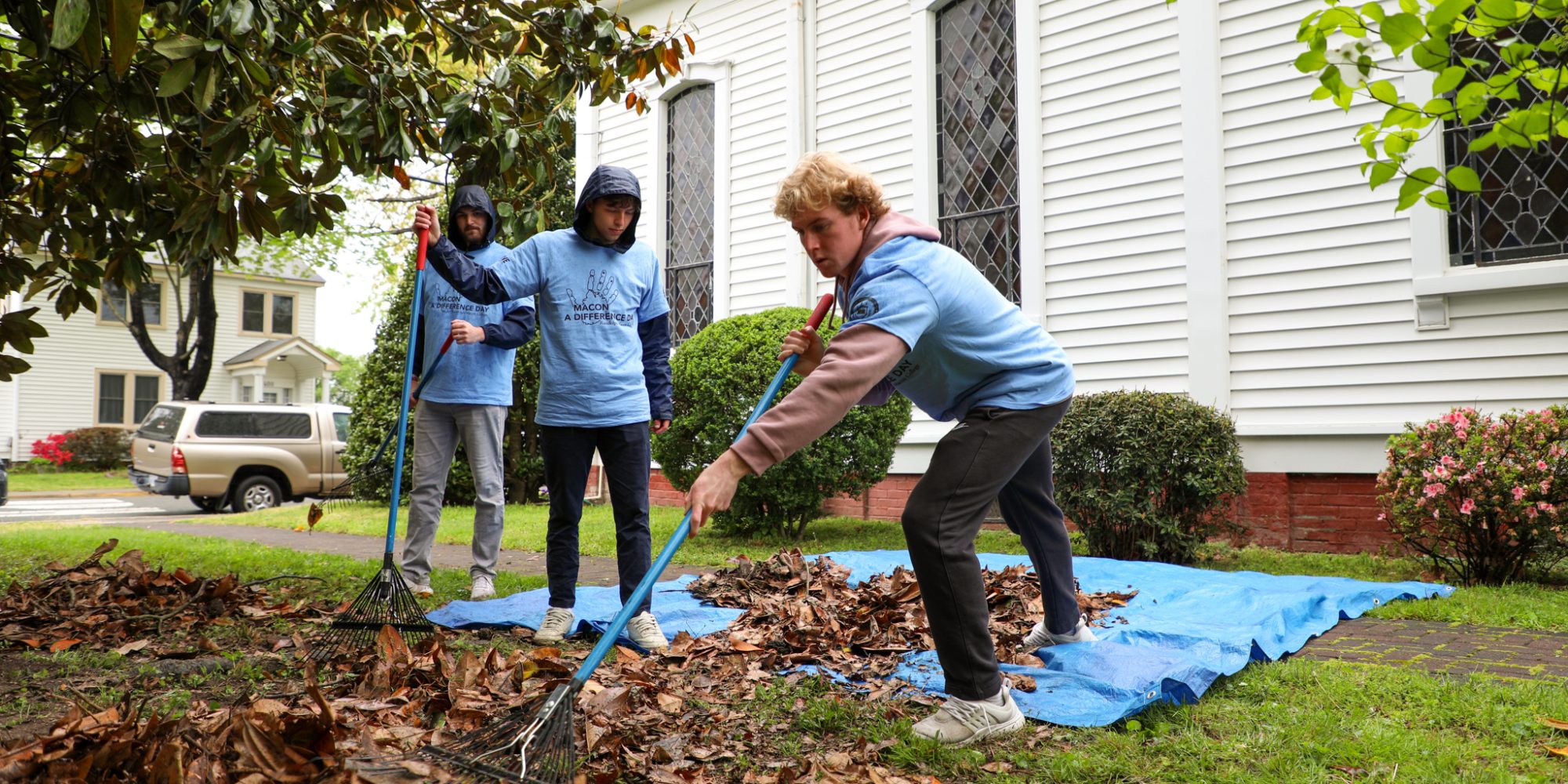 Three people in blue shirts, engaged in community service, rake leaves onto a blue tarp outside a white building.