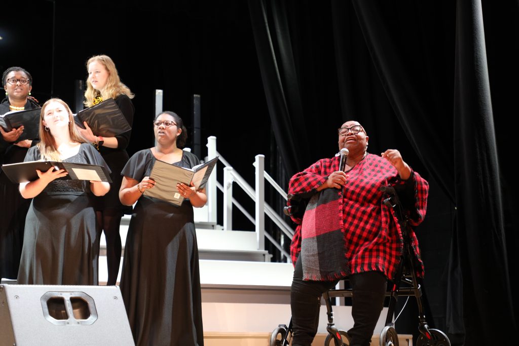 A group of performers in black outfits sings on stage. One person, wearing a red and black checkered outfit, holds a microphone and sits in a mobility aid.