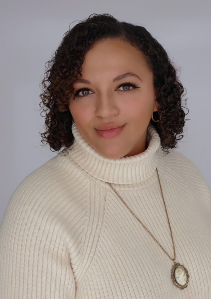 Person in a white turtleneck sweater with curly hair, wearing hoop earrings and a pendant necklace, smiles slightly at the camera.