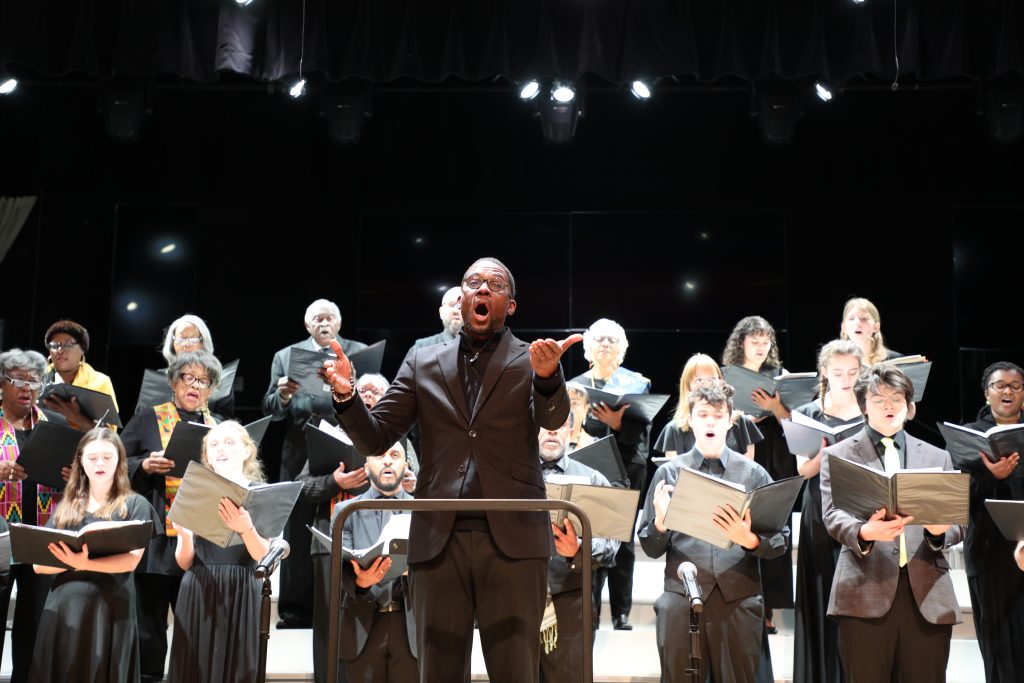 Conductor leading a choir on stage; members hold sheet music, singing under bright stage lights.