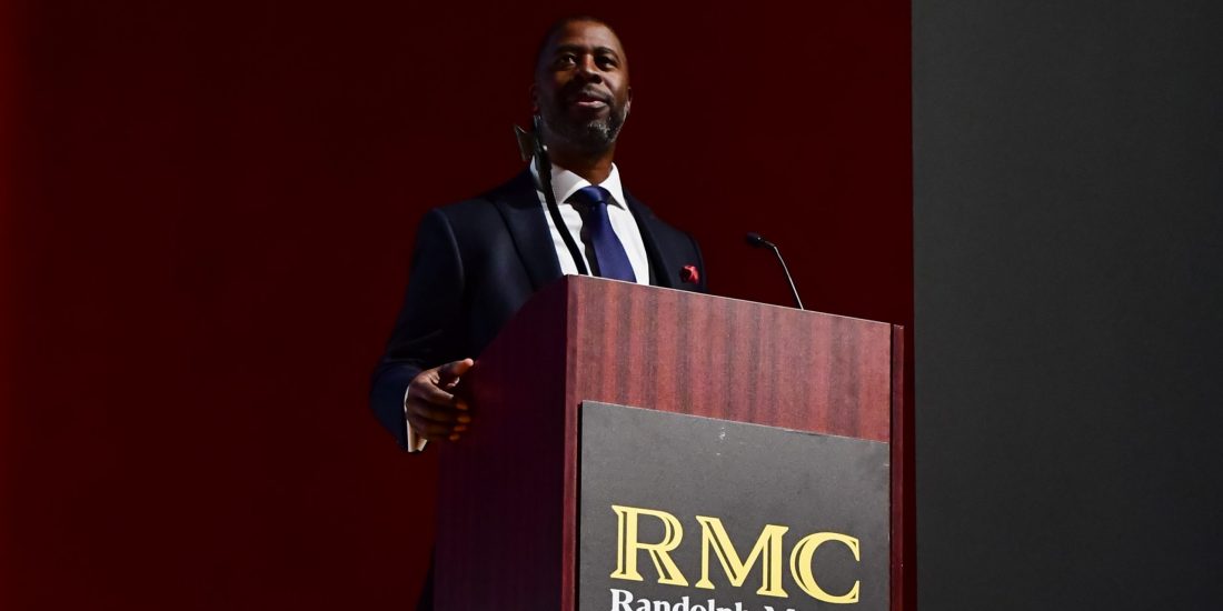 A man in a suit speaks at a podium labeled "RMC Randolph-Macon College" against a dark background.