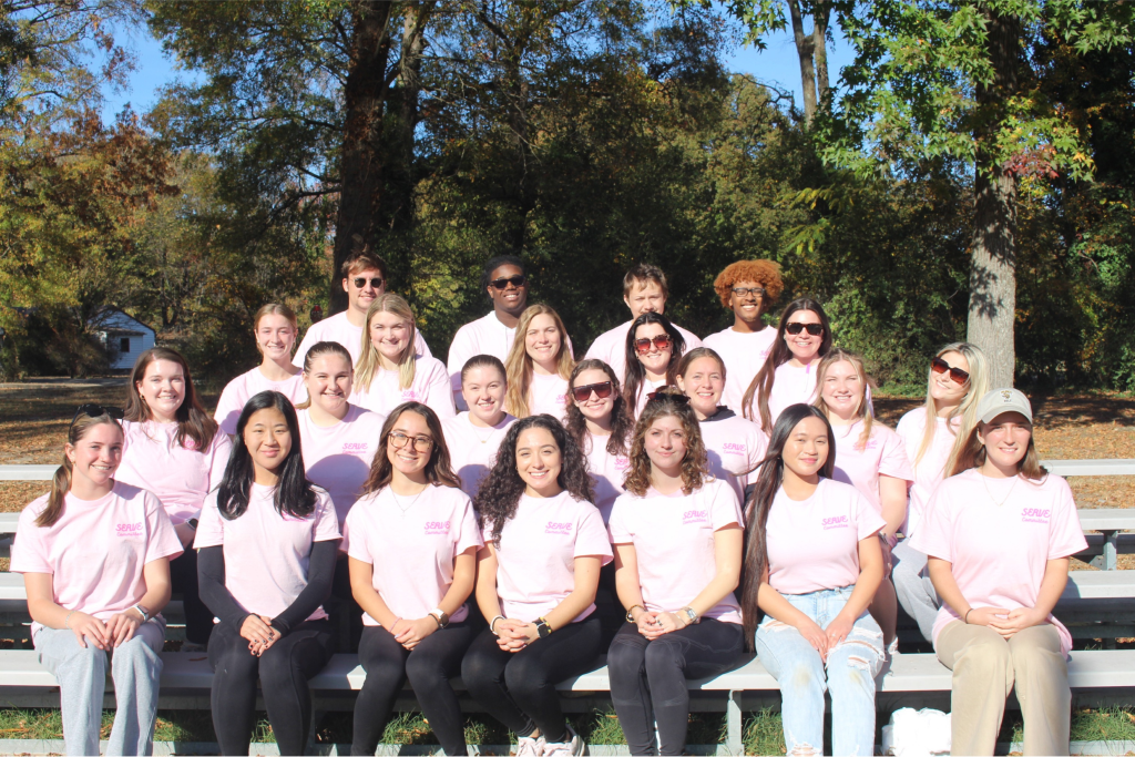 A vibrant community in pink shirts sits and stands on bleachers outdoors, surrounded by lush trees, ready to offer service together.