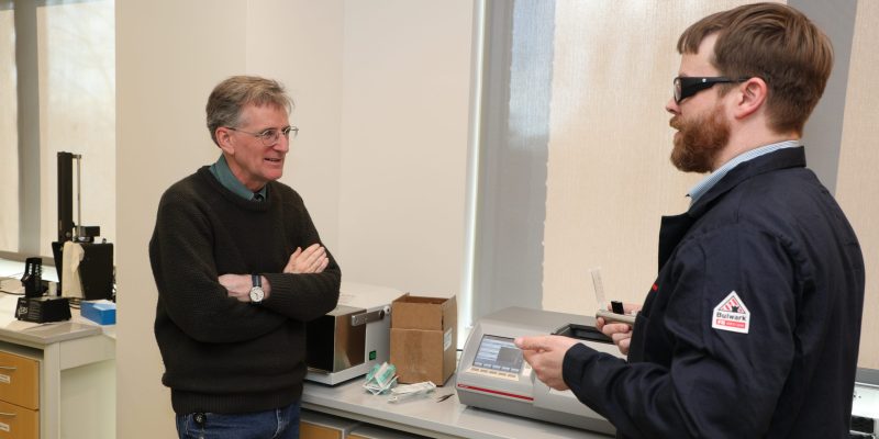 Two men talk in a laboratory. One holds a sample to be placed in a polarimeter; the other has arms crossed. Lab equipment is visible around them.
