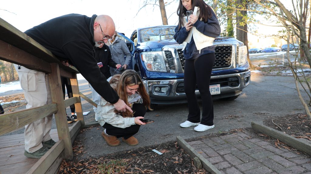 A small group of people observe something on the ground near a bridge. A blue truck is parked in the background.