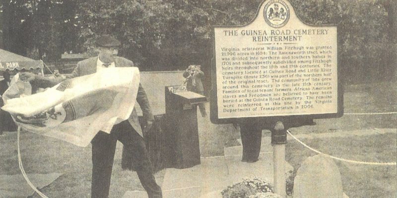 A person unveils a historical marker about the reinterment at the Guinea Road Cemetery, with a crowd observing in the background.