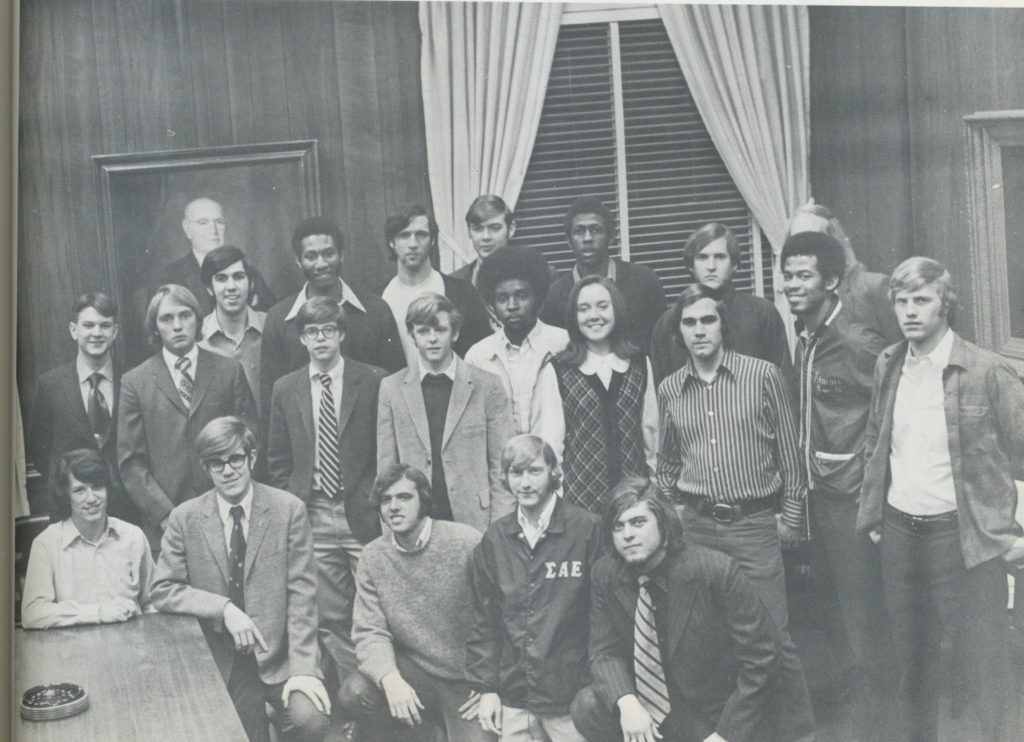 Black and white photo of a group of people in formal and semi-formal clothing, posing indoors with curtains and a portrait in the background.