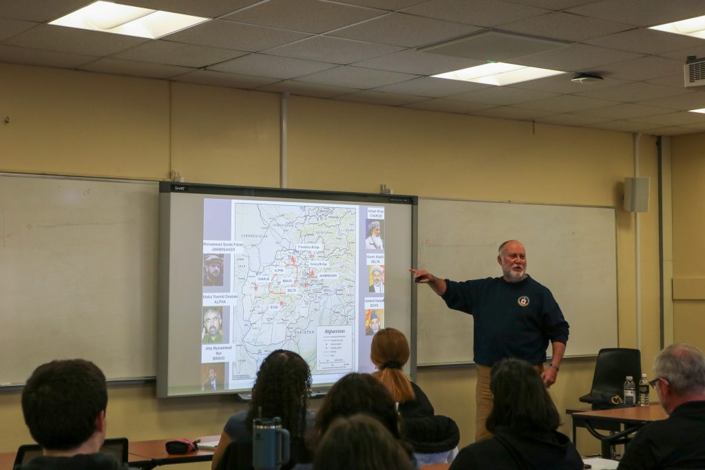 A bearded man giving a lecture, pointing at a map of Afghanistan projected on a screen, with an audience seated in front of him.