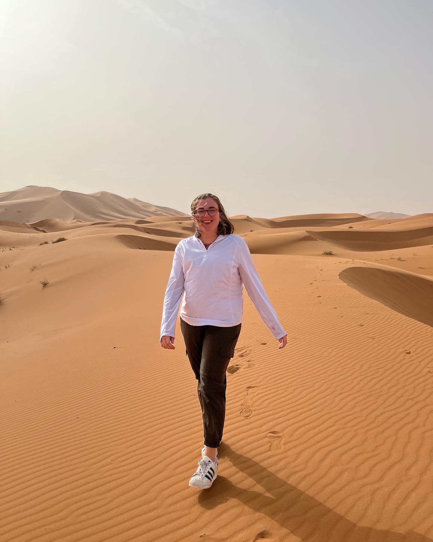 Matty Grace Gilliam during her Stud Abroad semester in Morocco strolls across sand dunes under a clear sky, wearing a white shirt and dark pants.