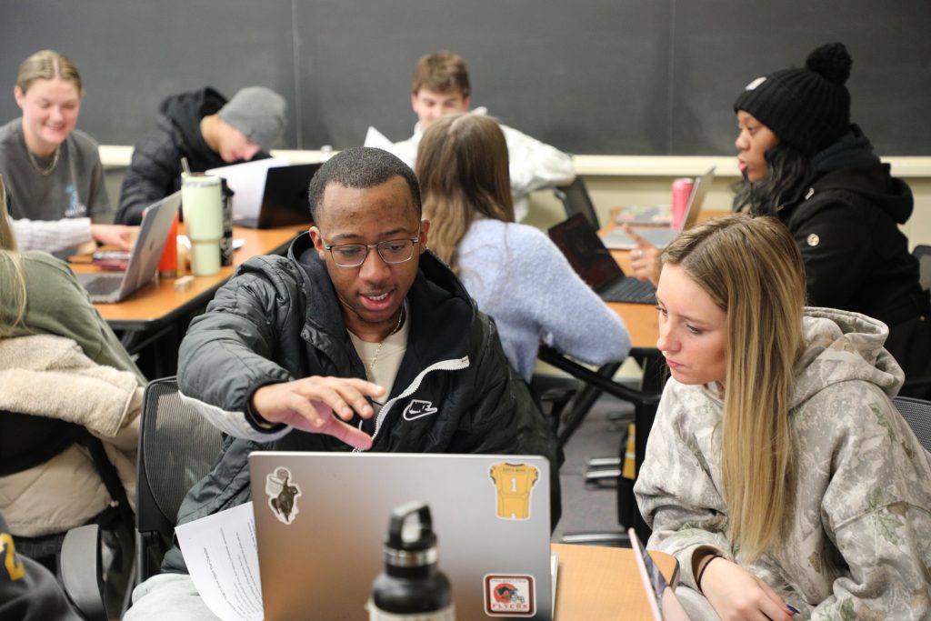 Students in a classroom engage in group work, sitting at tables with laptops and papers, discussing and collaborating in front of a chalkboard.