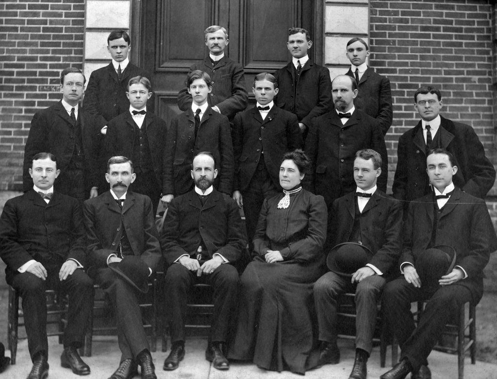 A vintage black and white photograph of fourteen people, mostly men in suits, with one woman, posed in front of a brick building.