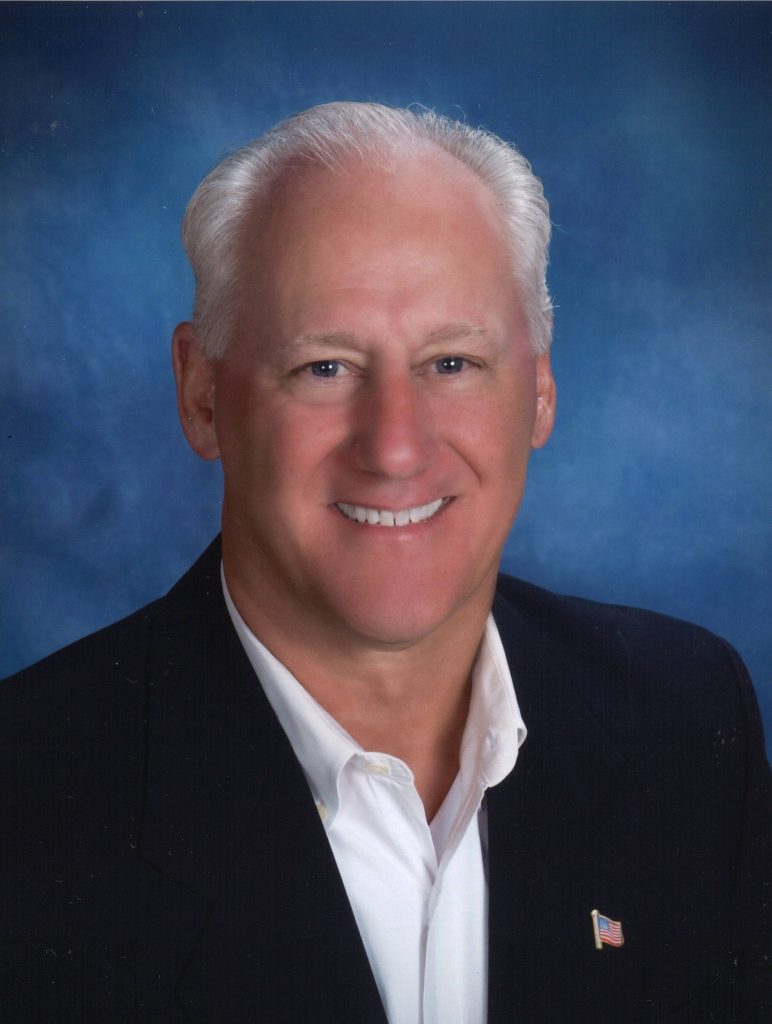 A smiling individual with white hair wearing a dark suit and white shirt is in front of a blue background. A small American flag pin is on the suit jacket.