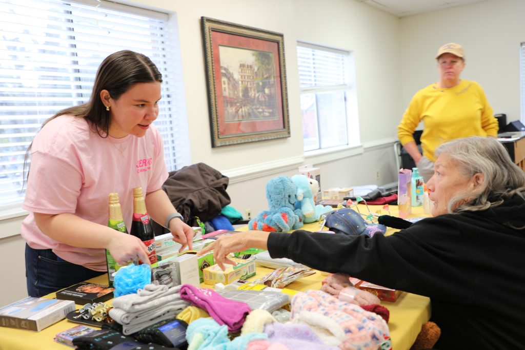 Woman in pink shirt assists an older woman selecting items from a table filled with various goods, including toys and toiletries. Another person in a yellow shirt stands in the background.
